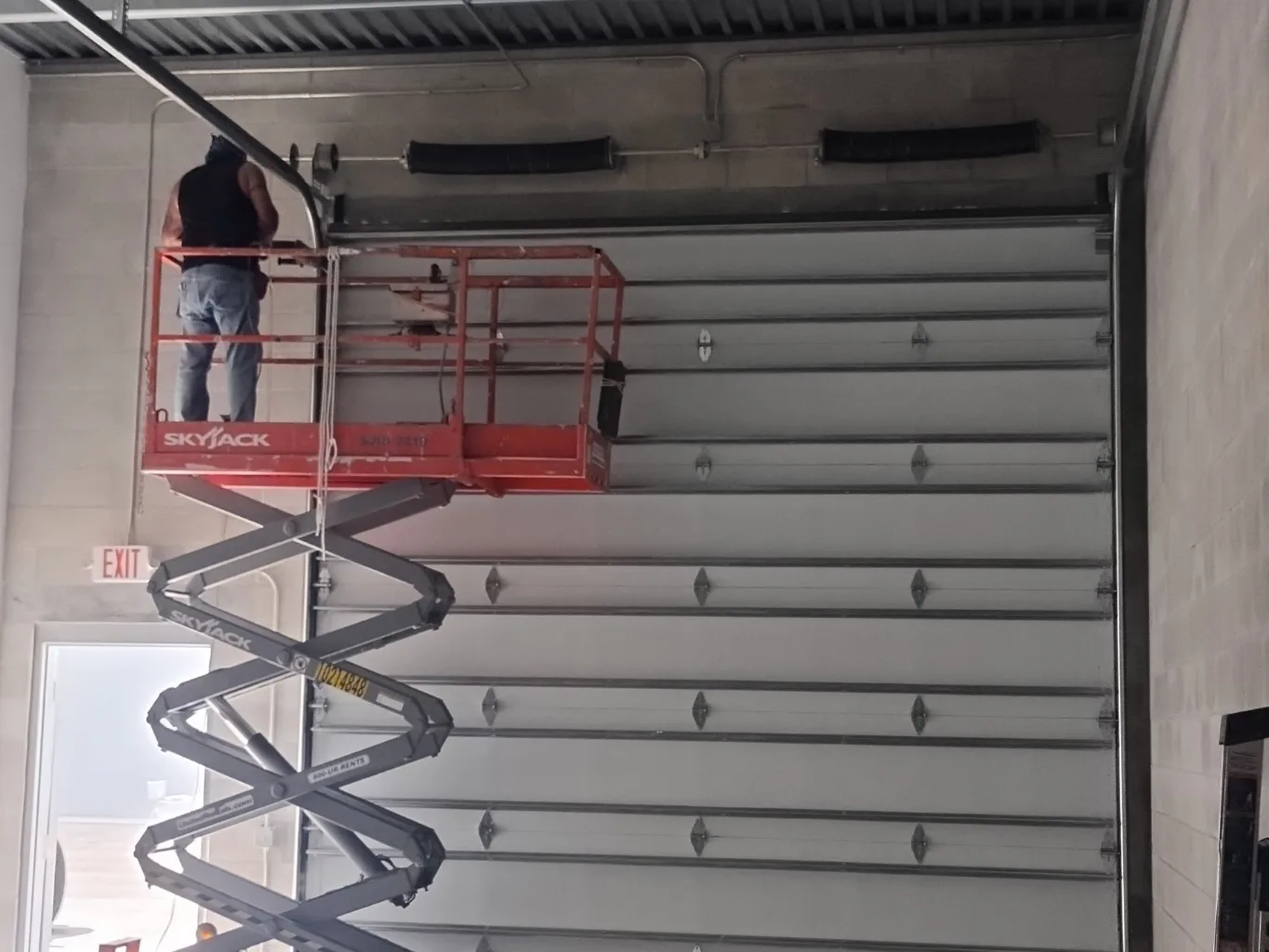 A garage door technician working on a residential safety inspection job — mid-task, hands, tools and the work itself in sharp focus, the rest of the space softly out of focus